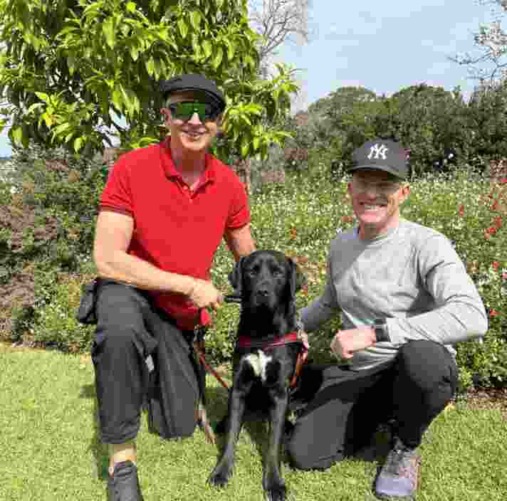 Scott with his black Labrador Guide Dog Pretzel and his partner Geoffry smiling at the camera.
