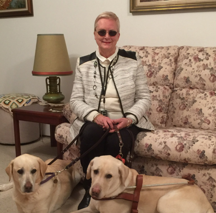 A woman sits on a couch with two Labradors at her feet.