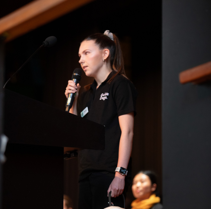 Lived Experience Presenter, Karlee, speaks into a microphone with her Guide Dog beside her.