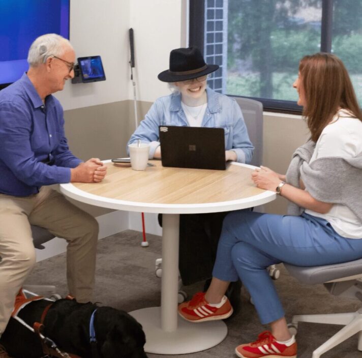 Three people sitting at a desk with a Guide Dog and a white cane,