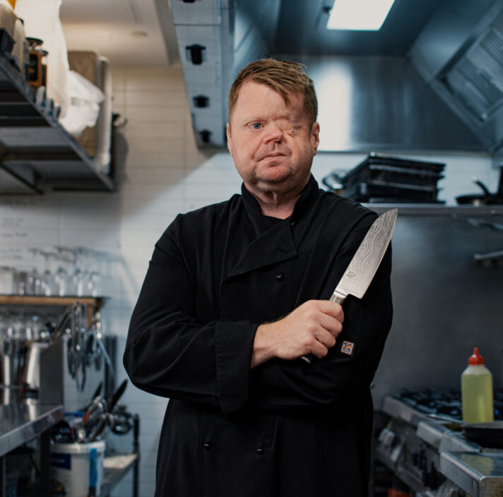 Craig stands in a commercial kitchen holding a large chef's knife.