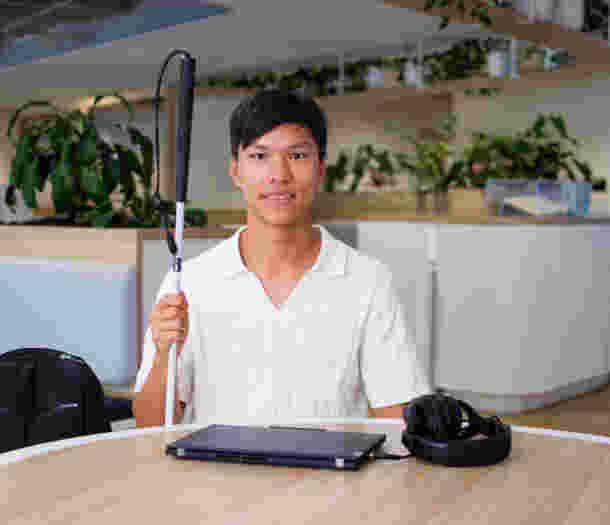 Seth sits at round table holding his white cane with a laptop in front of him.
