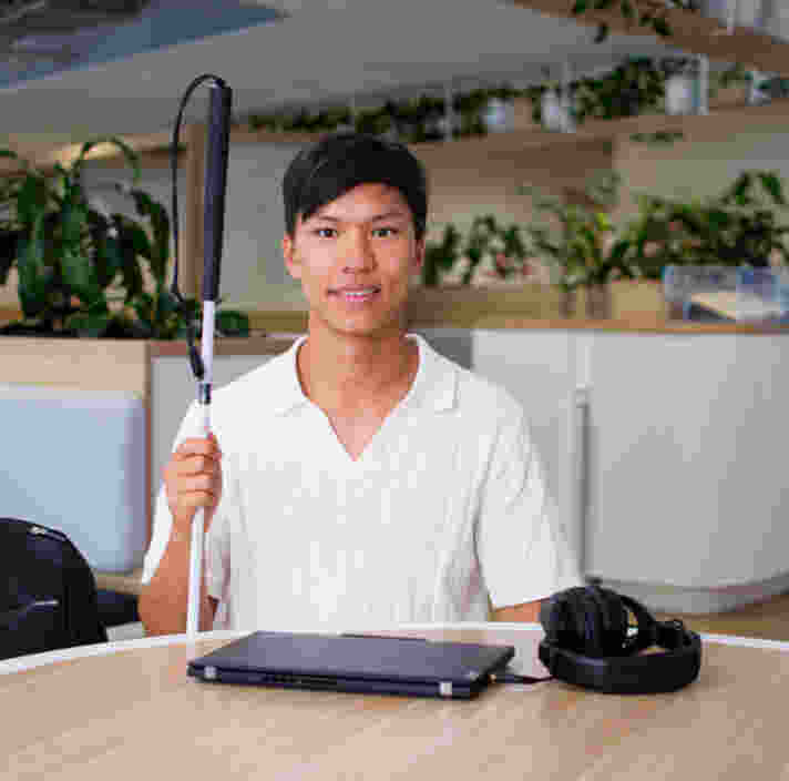 Seth sits at round table holding his white cane with a laptop in front of him.