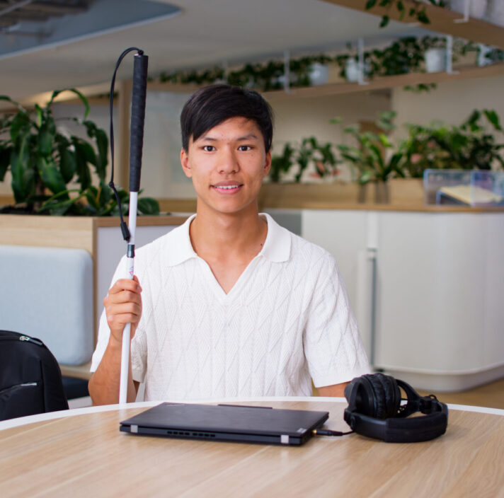 Seth sits at round table holding his white cane with a laptop in front of him.