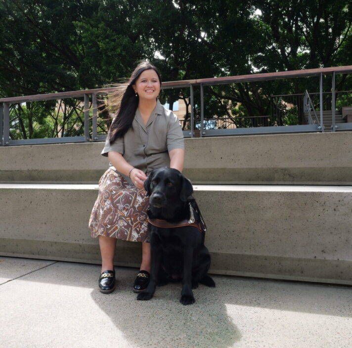 Ria and black Labrador guide dog sitting down outside.