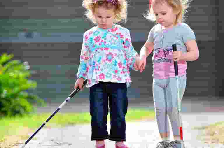 Two young girls, one holding a white cane, walk hand in hand on a brick path.
