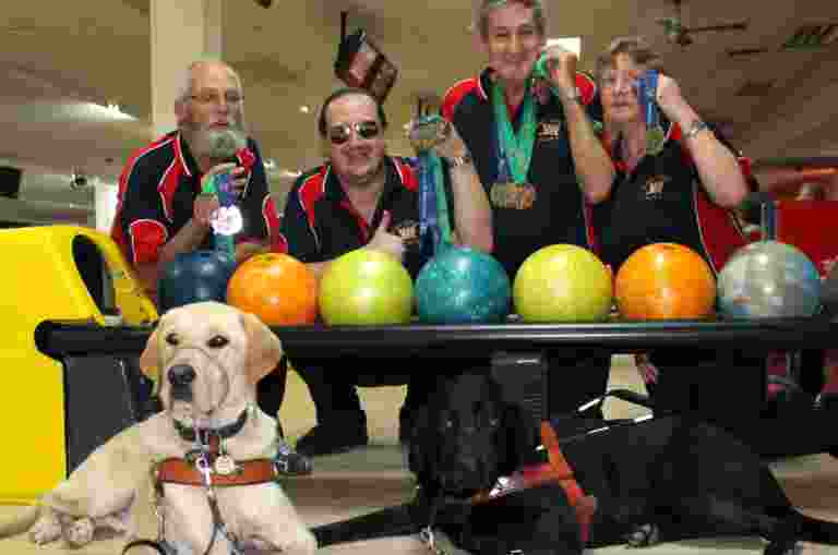 A group of four bowlers hold their gold and silver medals. They are inside a bowling alley with two Guide Dogs.