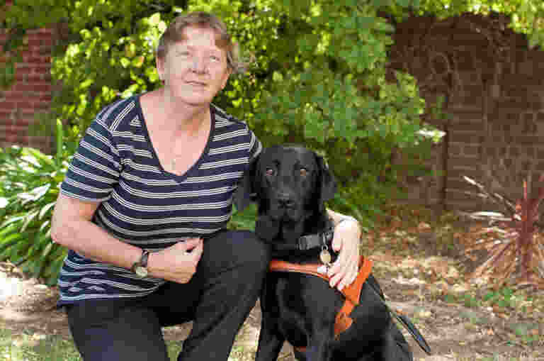 Marie kneels beside her black Guide Dog, Ebony, in a leafy garden