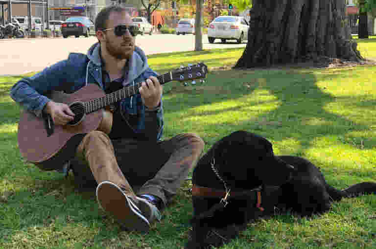 A man, Arron, sitting outside on the grass playing the guitar with a Black Guide Dog, Helix, laying beside him