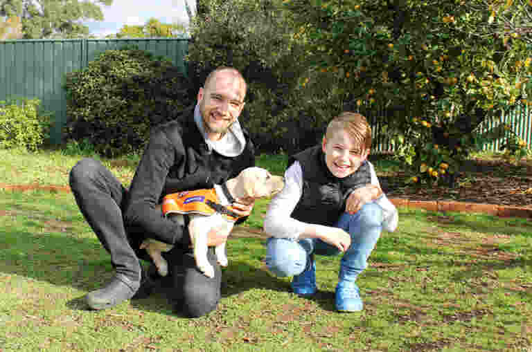 A man holding a small Yellow Labrador in an orange training jacket kneeling outside on the ground next to a young boy