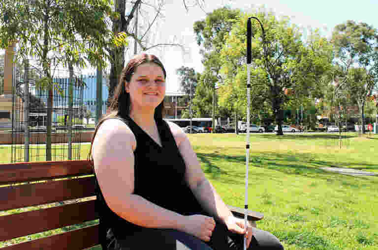 A woman sitting outside on a park bench holding a white cane and smiling at the camera
