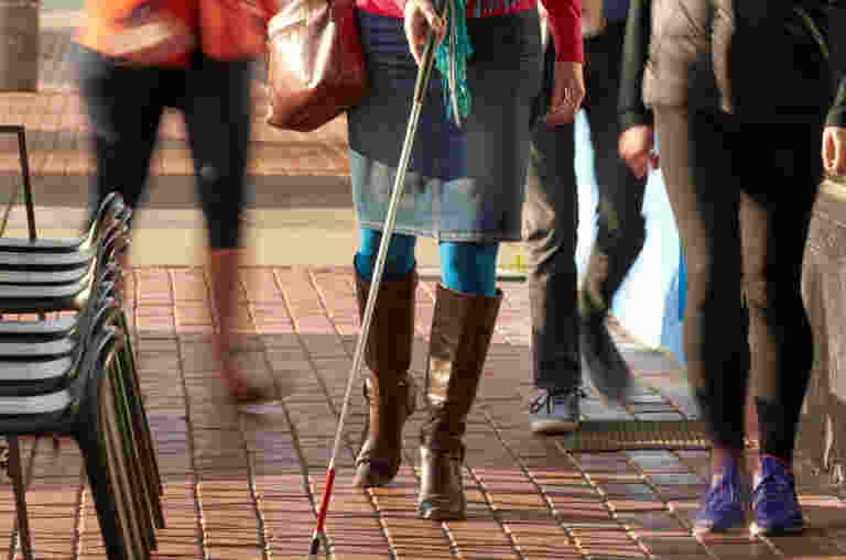 A woman with a white cane is walking along a busy footpath