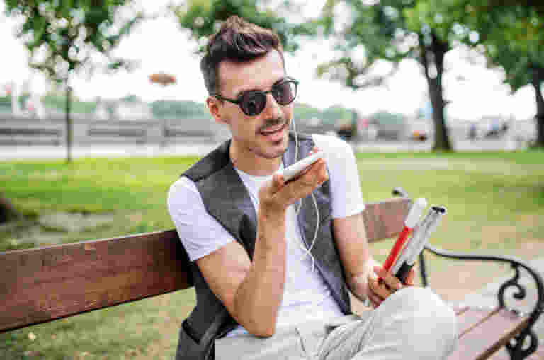 A person sitting outside on a park bench wearing sunglasses and headphones, holding a cane and a phone in their hands. The person is talking into the phone.