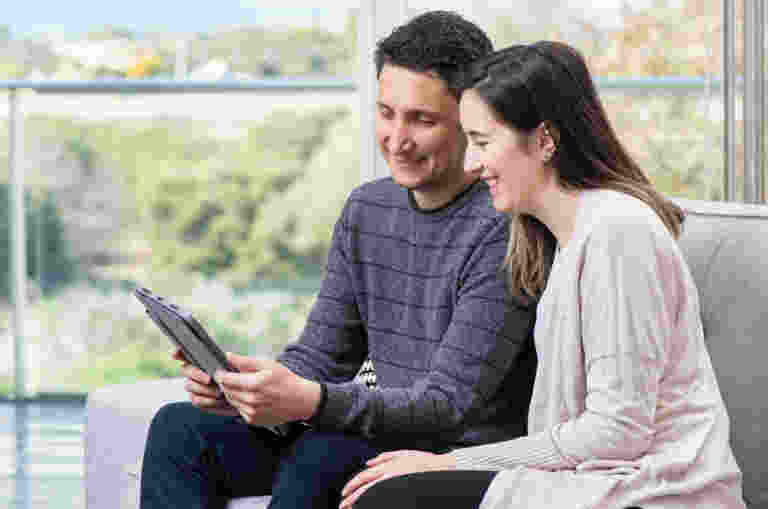 A man and woman sitting inside on a couch looking at a tablet computer