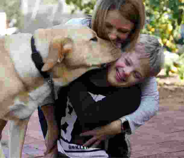A young child is getting a kiss from a yellow labrador dog. His mother is behind them giving them a cuddle.