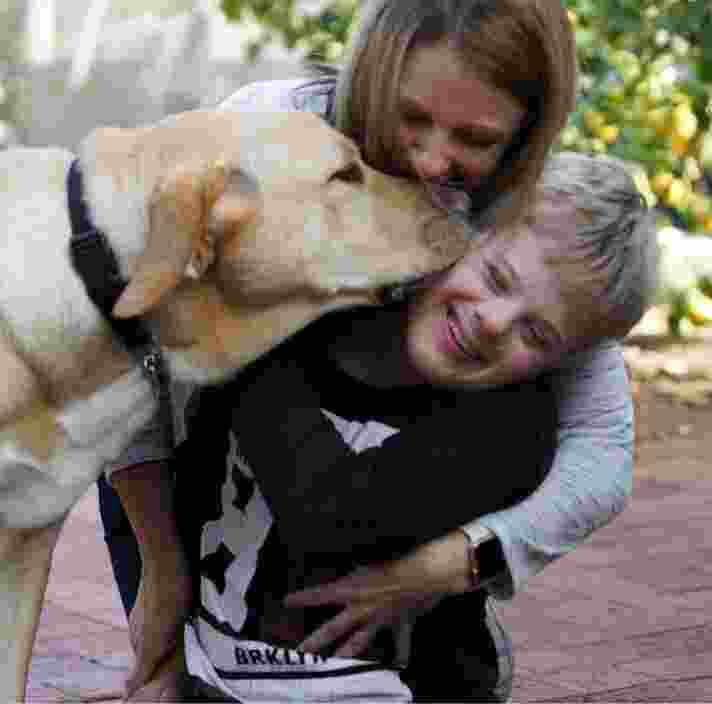A young child is getting a kiss from a yellow labrador dog. His mother is behind them giving them a cuddle.