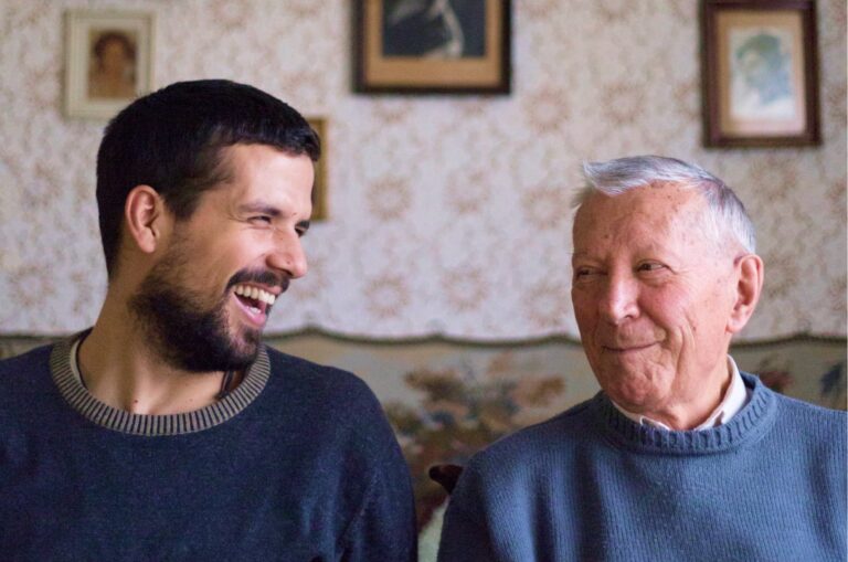 Two people, one adult & one older person, sitting on a couch looking at each other laughing.