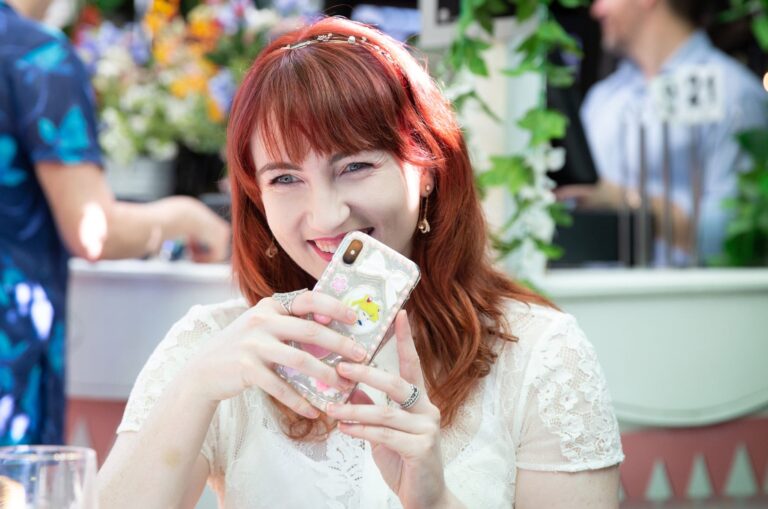 An adult who is outside at a cafe laughing. They have their phone up to their mouth and it is covering part of their mouth.
