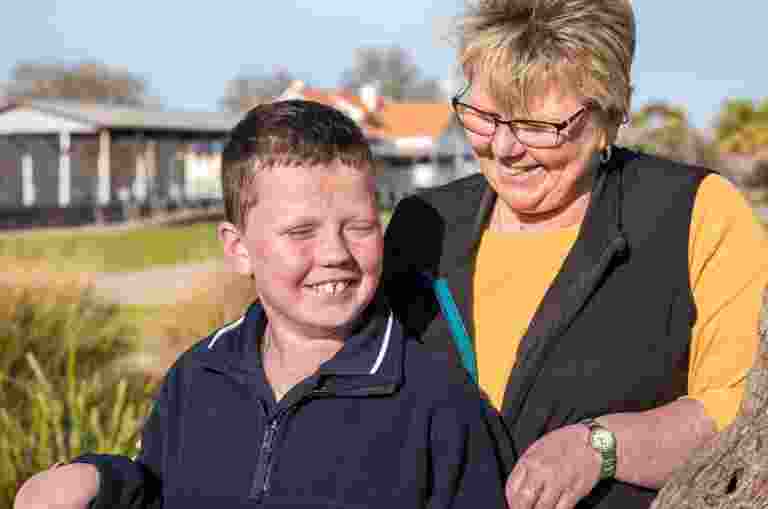 A young boy standing outside with his mother. The mother is close behind her son and they are both smiling, The mother is looking at her son.