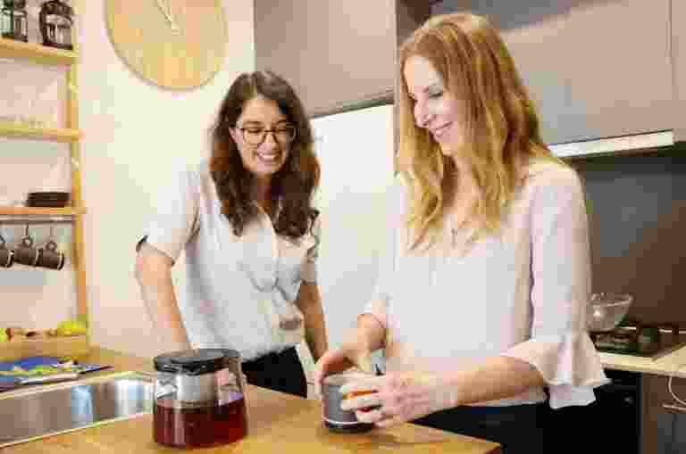 Two people inside in the kitchen. One is making tea while the other is watching on.