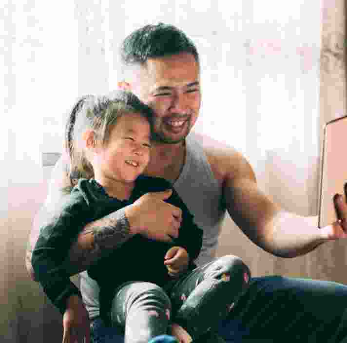 A young child sitting on their father's lap while they both smile while looking at an Ipad. The father has one had around his child and is holding the Ipad with the other hand.