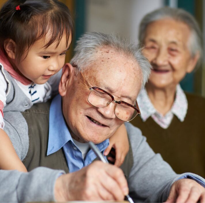 An older man writing while a young girl puts her arms around his shoulders, hugging him. The man is smiling and there is an older woman in the background who is not in focus but is also smiling.