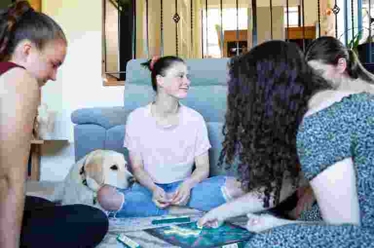 A group of four young adults sitting inside, in a circle. There is a yellow labrador dog who is resting its head on one of the person's legs.