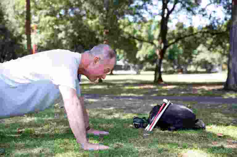 A person doing push ups outside in the park. There is a black bag near them with a folded white cane leaning up against the bag.