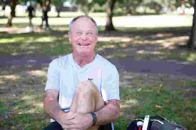A person sitting outside on the grass with a smile on their face looking at the camera. They have one leg bent with their arm wrapped around their knee and a white cane next to them.