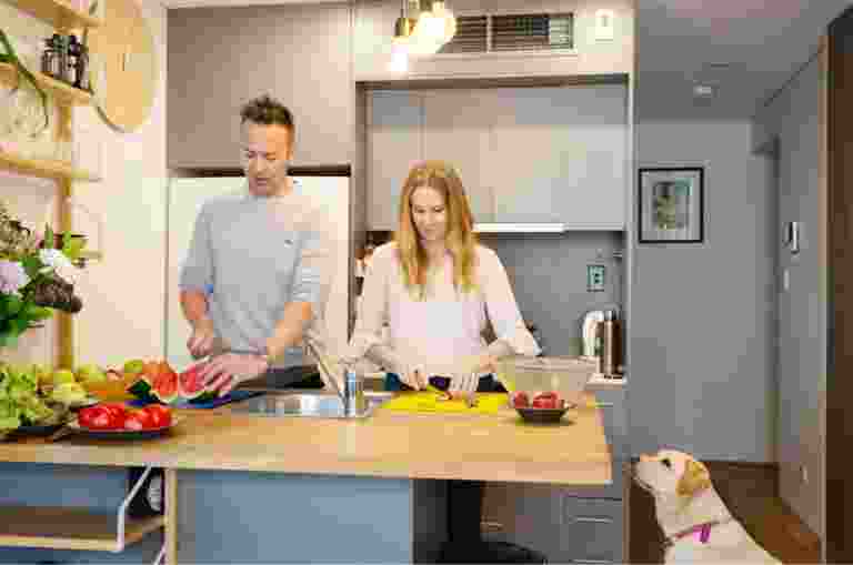 A couple preparing fresh food in their kitchen. Next to the kitchen table is a yellow labrador dog sitting on its back legs looking up at the food.
