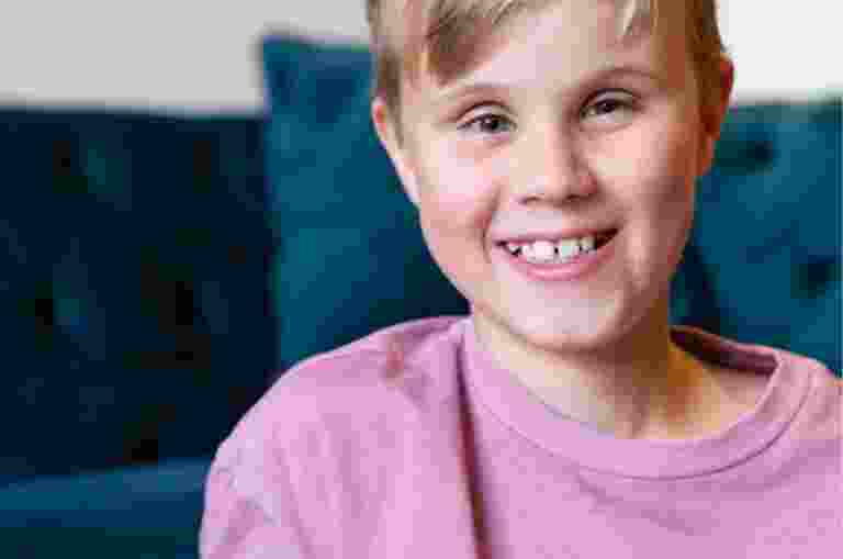 Close up of a young child with a big smile on their face. They are seated on the ground with a couch in the background.