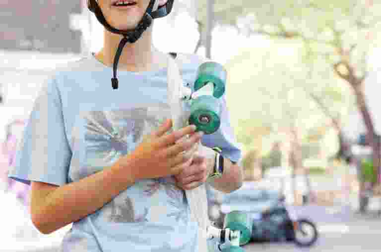 Young child wearing a helmet, holding a skateboard and standing outside in the city streets. The child is looking at the camera and smiling.