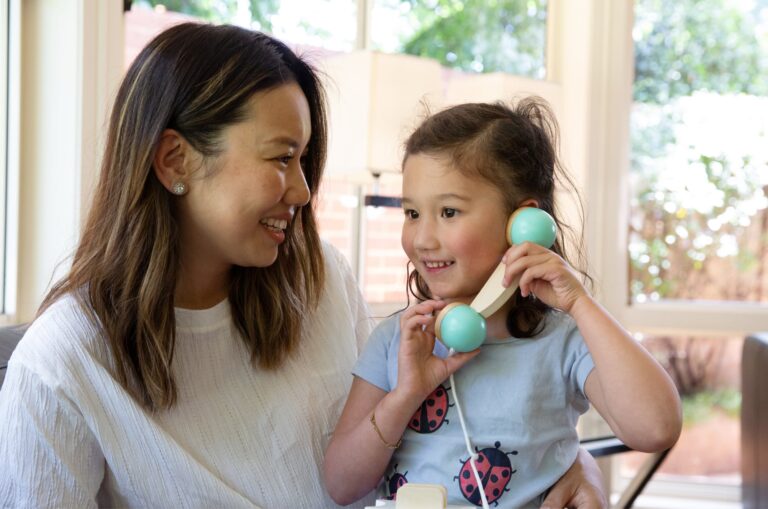 A child plays with a pretend phone while a mother sits next to her smiling.