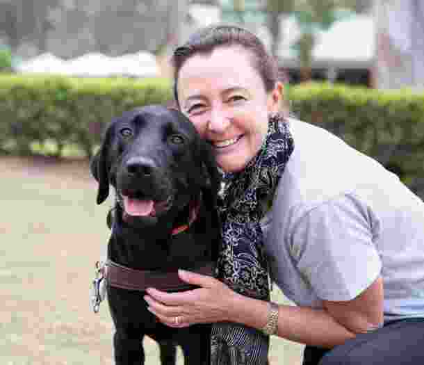 A person hugging a black labrador Guide Dog in harness. The person and dog are looking at the camera and the person is smiling,