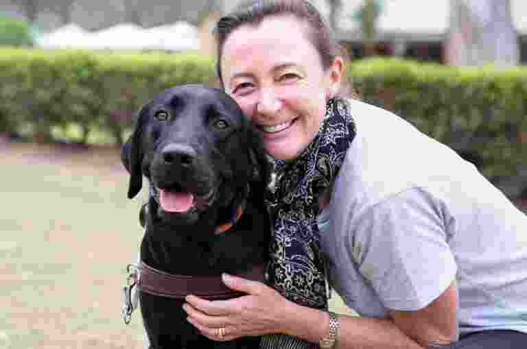 A person hugging a black labrador Guide Dog in harness. The person and dog are looking at the camera and the person is smiling,