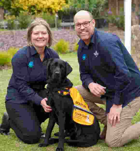 Two people on either side of a black Autism Assistance Dog. The people are kneeling down next to the dog and the photo is taken outside on grass. The dog is wearing an Autism Assistance Dog coat and everyone is looking at the camera.