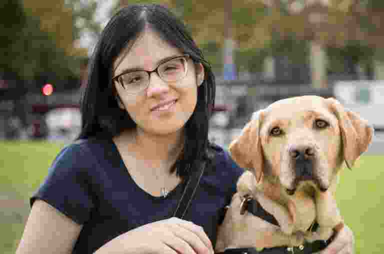 A person with glasses kneels on grass beside a yellow Labrador wearing a harness.