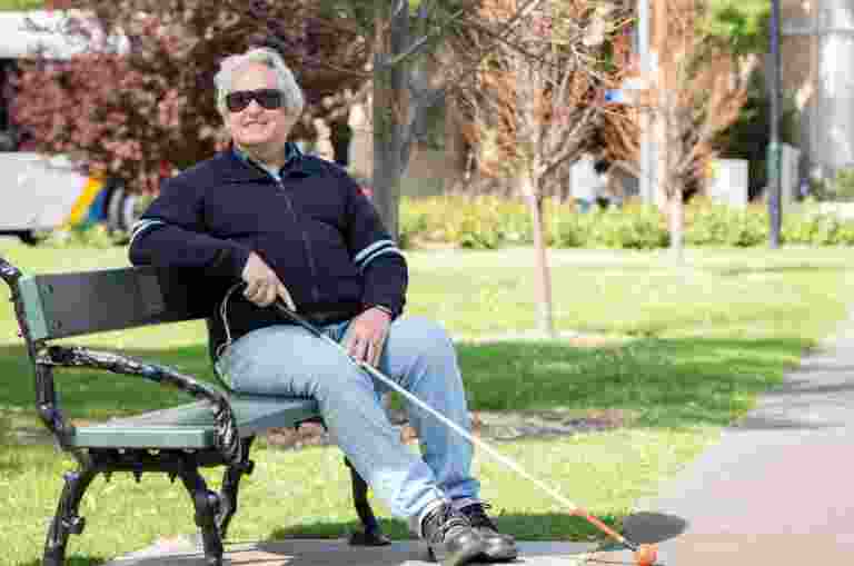 A person sitting on a park bench, holding a white cane with a red tip, wearing sunglasses and a dark jacket.