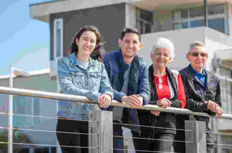 Four people standing outdoors, leaning on a railing, with a modern building in the background.