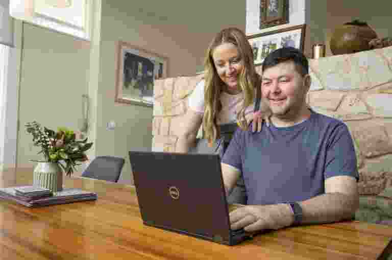 A person sitting at a table looking at a laptop with another person leaning on his shoulder, also looking at the laptop.