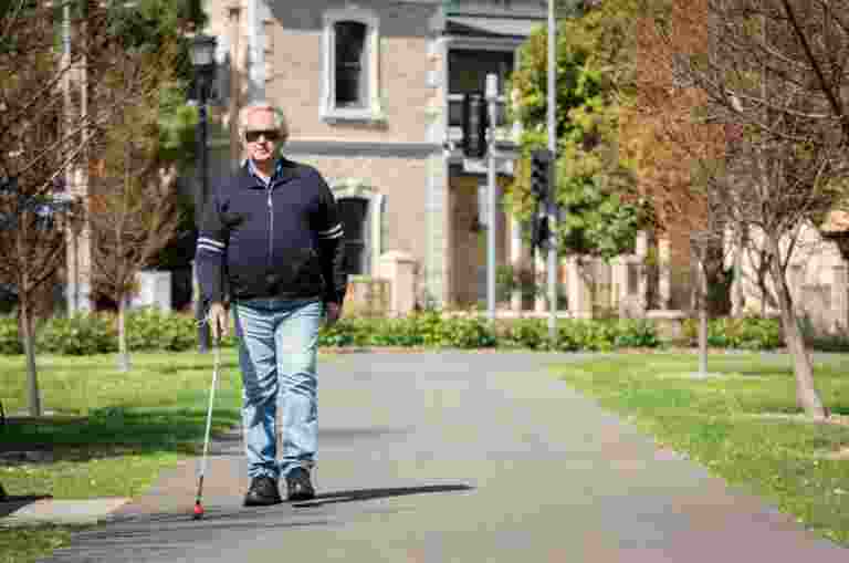 A man with a white cane walks on a park pathway, surrounded by green grass and trees, with a stone building in the background.