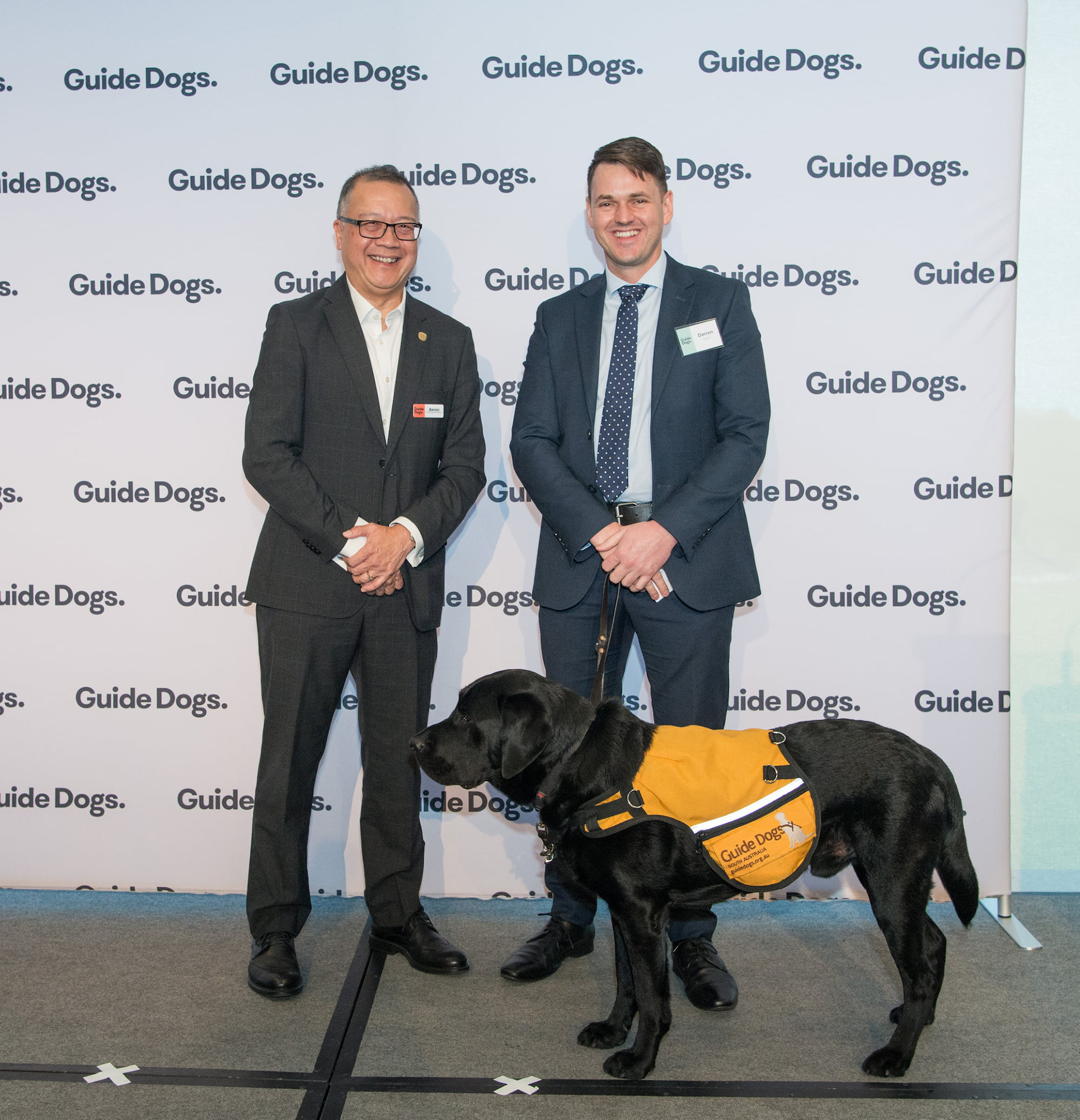 Guide Dogs SA/NT CEO Aaron Chia is standing on stage next to Canine Court Companion Zero, a black Labrador, and his handler Darren.