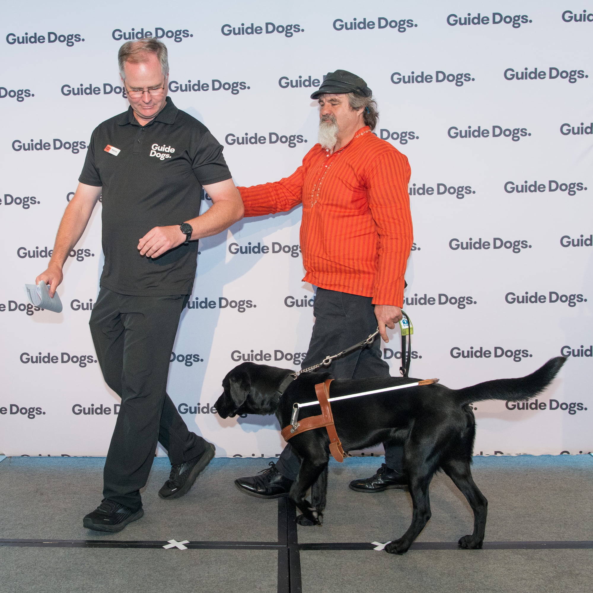 Guide Dogs Client Anthony with his Guide Dog Kit, a black Labrador wearing a harness, being led on stage by a Guide Dogs staff member.