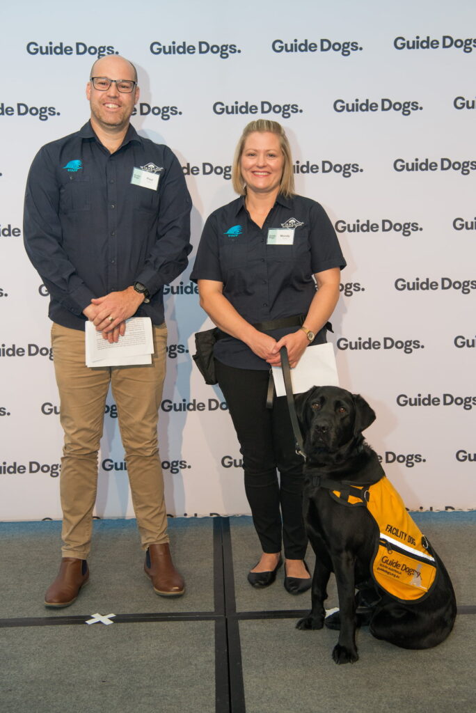 Iris, a black Labrador wearing a yellow Facility Dog jacket, standing on stage with her handlers Paul and Mandy.