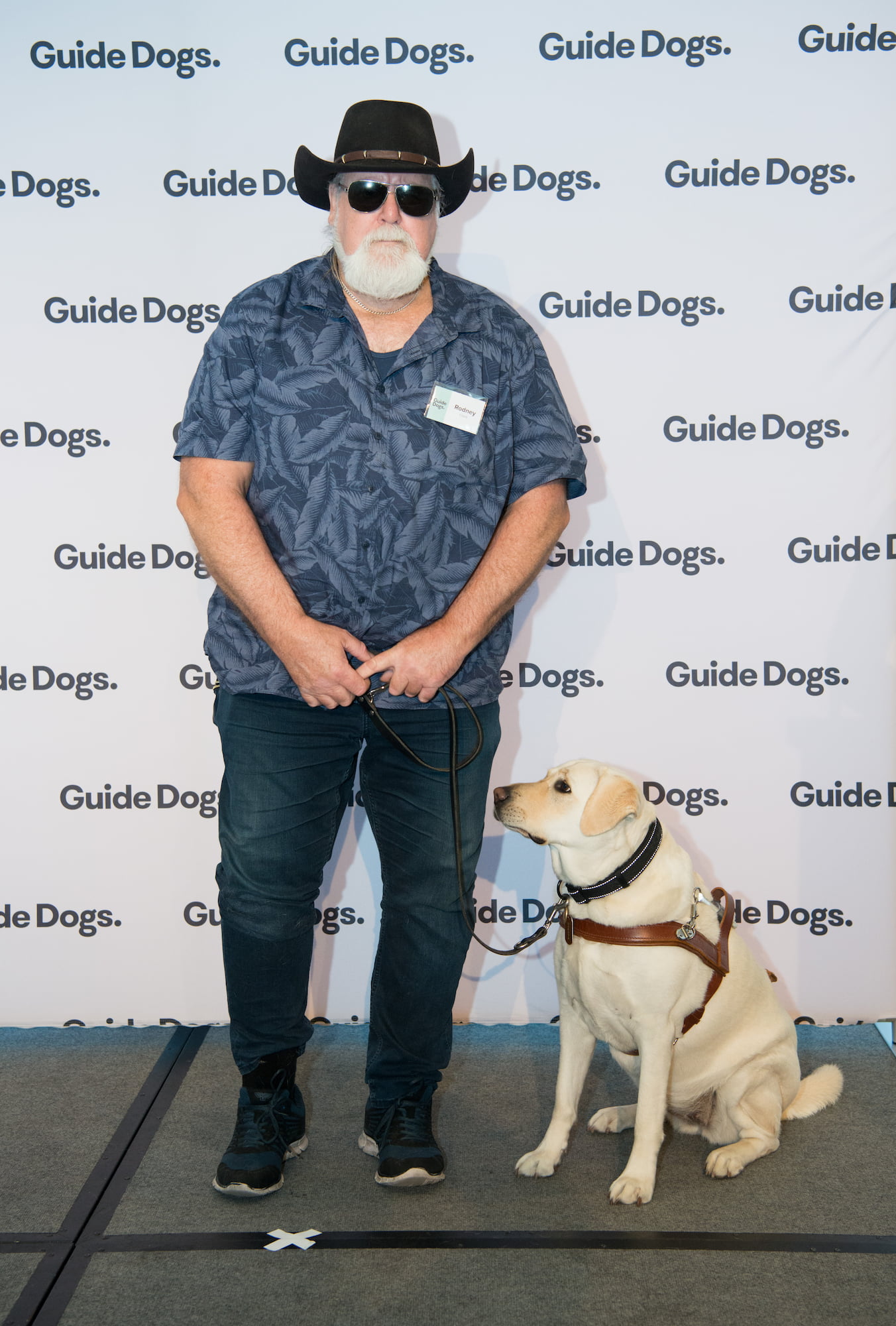 Guide Dogs Client Rodney standing on stage with his Guide Dog Kit, a yellow Labrador wearing a harness.