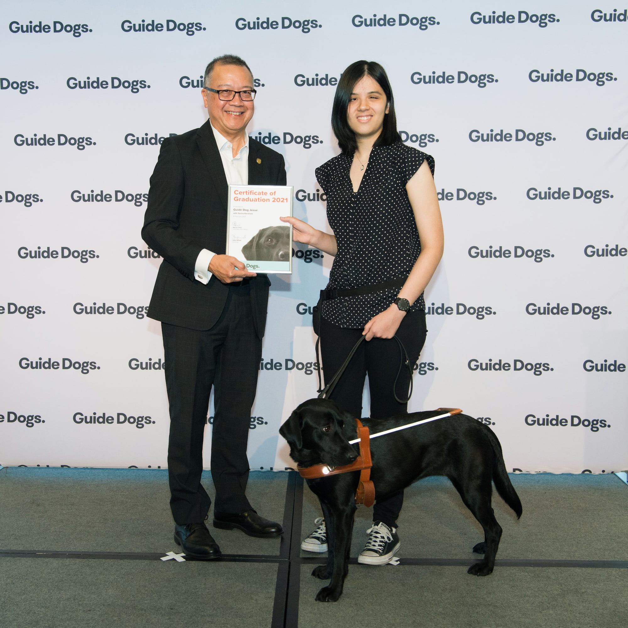 Guide Dogs SA/NT CEO Aaron Chia standing on stage with Guide Dogs Client Sacha and her Guide Dog Jesse, a black Labrador who is wearing a harness. Aaron is presenting Sacha with a certificate.