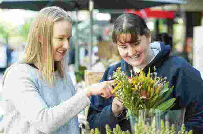 Two women smiling and looking at a bouquet of flowers at an outdoor market.