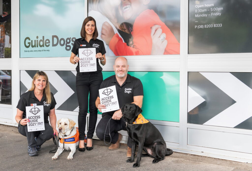 Three Guide Dogs staff members stand outside the Guide Dogs SA/NT building holding the 2021 Adelaide Fringe Access Guide. With them is two Labrador dogs.