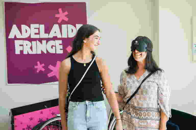 Two people are standing in front of an Adelaide Fringe sign. One of them is wearing a blind fold and holding on to the other person’s arm.
