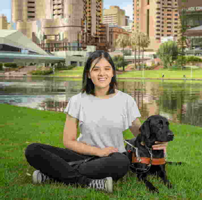 A person sitting on grass smiling at the camera. There is a Black Guide Dog in harness laying beside the person looking into the distance.
